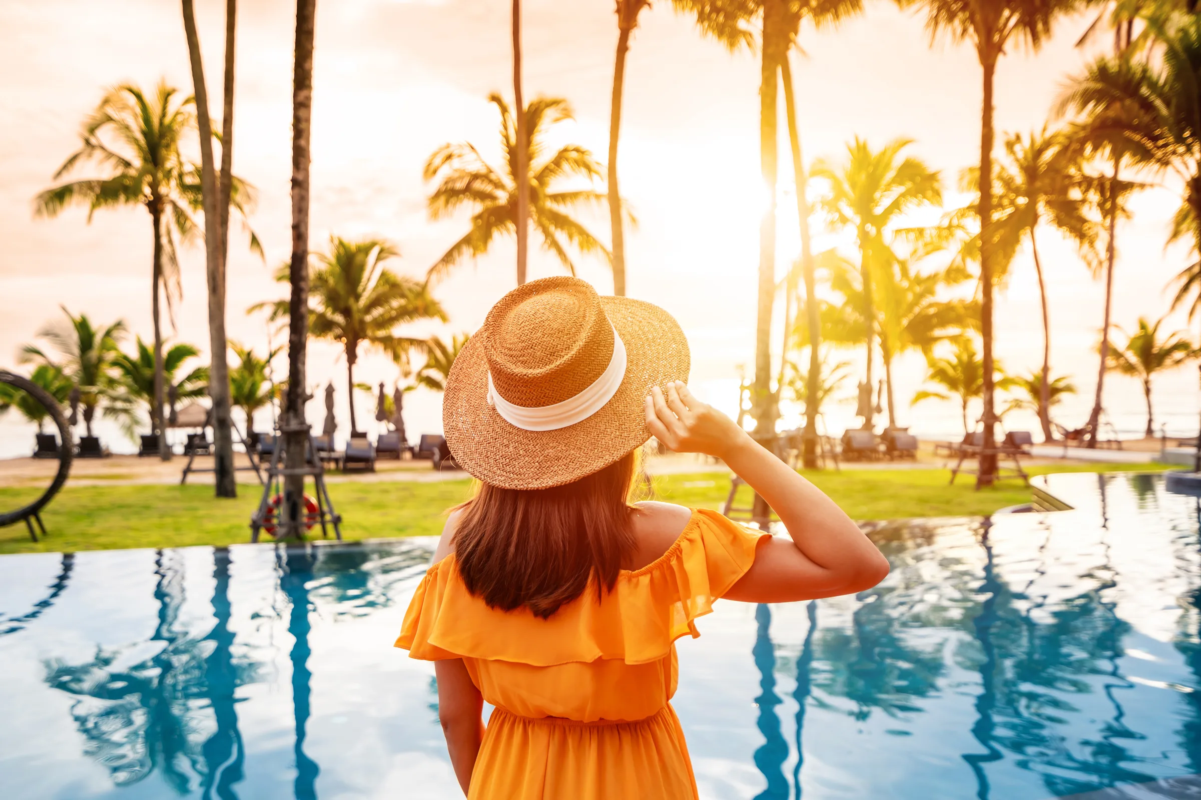 A guest in a sun hat by a tropical resort pool, framed by palms at golden hour