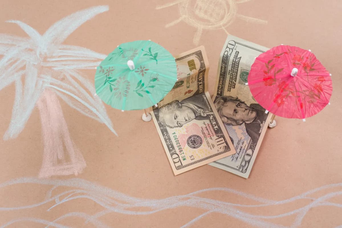 Ten and twenty dollar bills with paper cocktail umbrellas over sand-colored paper with chalk drawings of a palm tree and waves